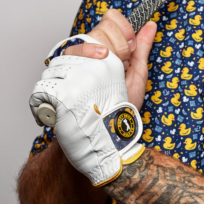 A tattooed arm grips a golf club while wearing the Big Duck Energy - Glove, a precision-fit cabretta leather golf glove with yellow trim and Birdie Bros Golf Co. logo. The person sports a navy shirt with yellow ducks and "BAE" text on it.