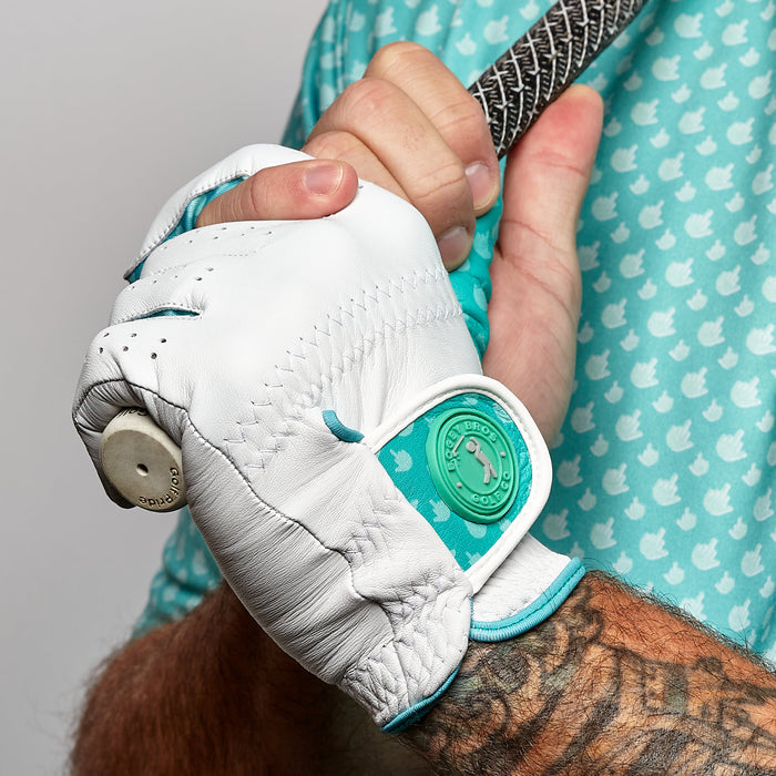 A close-up of a tattooed hand gripping a golf club while wearing the Birdie Finger - Glove, a precision fit cabretta leather glove with teal accents and round logo; paired with a teal, white bird-patterned shirt for stylish golfing.
