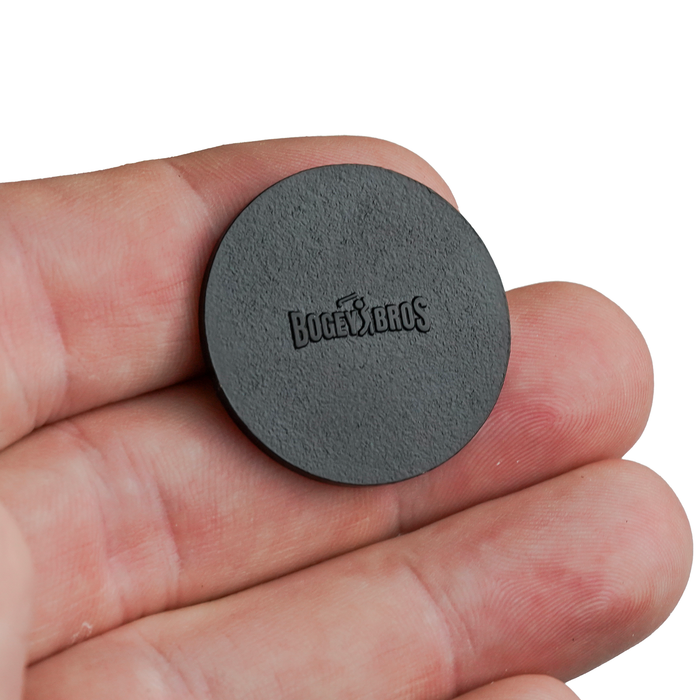A close-up of a hand holding the Bogeyman - Ball Marker, a small, round, black heavy metal marker with iron nickel plating and the “BOGEY BROS” logo engraved at the center, fitting easily between fingertips to show its compact size.
