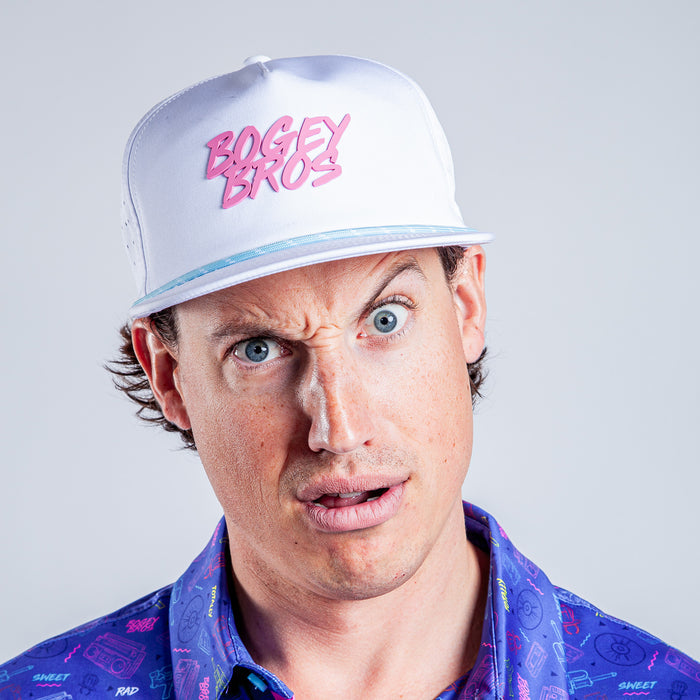 A man with light skin and blue eyes stares intensely, wearing the Da 90's - Unstructured Flat Bill Hat - Snapback with pink “BOGEY BROS” text and a blue patterned shirt against a light gray background.