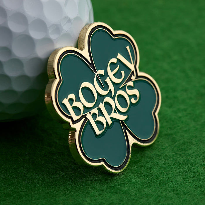 The Gettin' Lucky - Ball Marker, a gold and green four-leaf clover marked with BOGEY BROS in gold letters, rests on green turf beside a dimpled white golf ball.