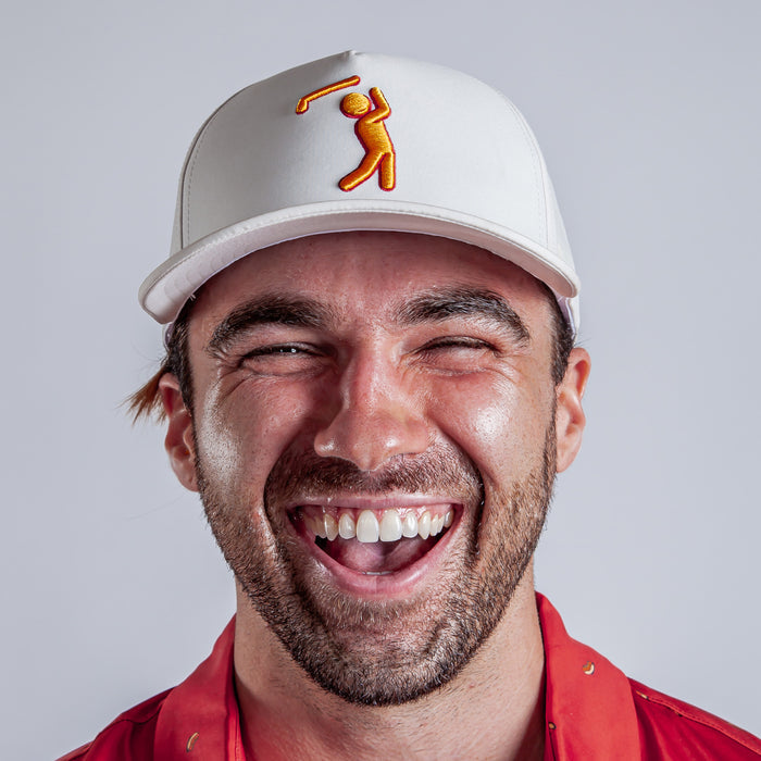 A bearded young man smiles widely, wearing the Glizz Reaper - Performance Golf Hat - Snapback, a white cap with an orange golf-swinging figure embroidered on it, paired with a red collared shirt. The background is plain light gray.