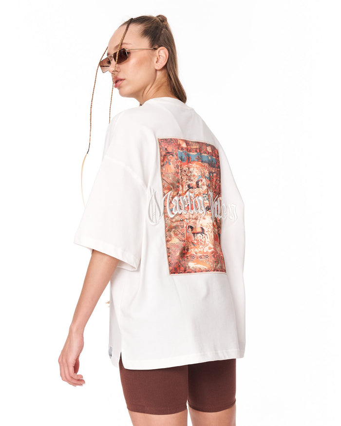 A woman wearing an oversized white short-sleeve t-shirt with a large multicolor graphic print on the back, paired with brown fitted shorts, photographed against a plain white background.