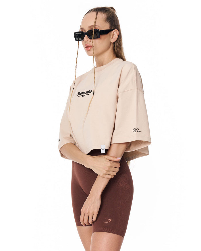 A model wearing a beige cropped t-shirt with black graphic text on the chest and a matching brown fitted biker shorts, styled with black sunglasses against a plain white background.
