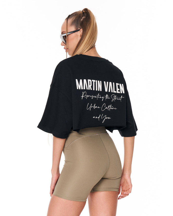 A woman shown from the back wearing a black oversized crop top with white text reading 'MARTIN VALEN' and beige fitted shorts against a plain white background.