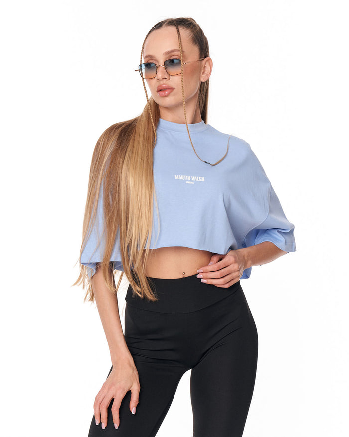 A woman modeling a light blue mock-neck crop top with 'MARTIN VALEN' printed in white on the chest, paired with black leggings, against a plain white background.