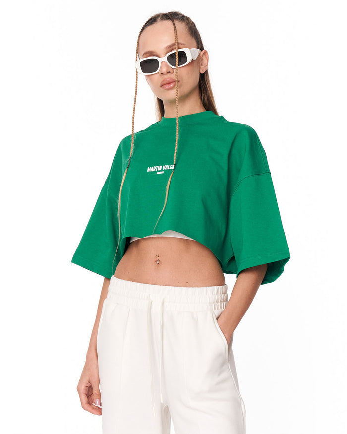 A model wearing a bright green cropped t-shirt with white text on the chest, paired with white sweatpants, against a plain white background.
