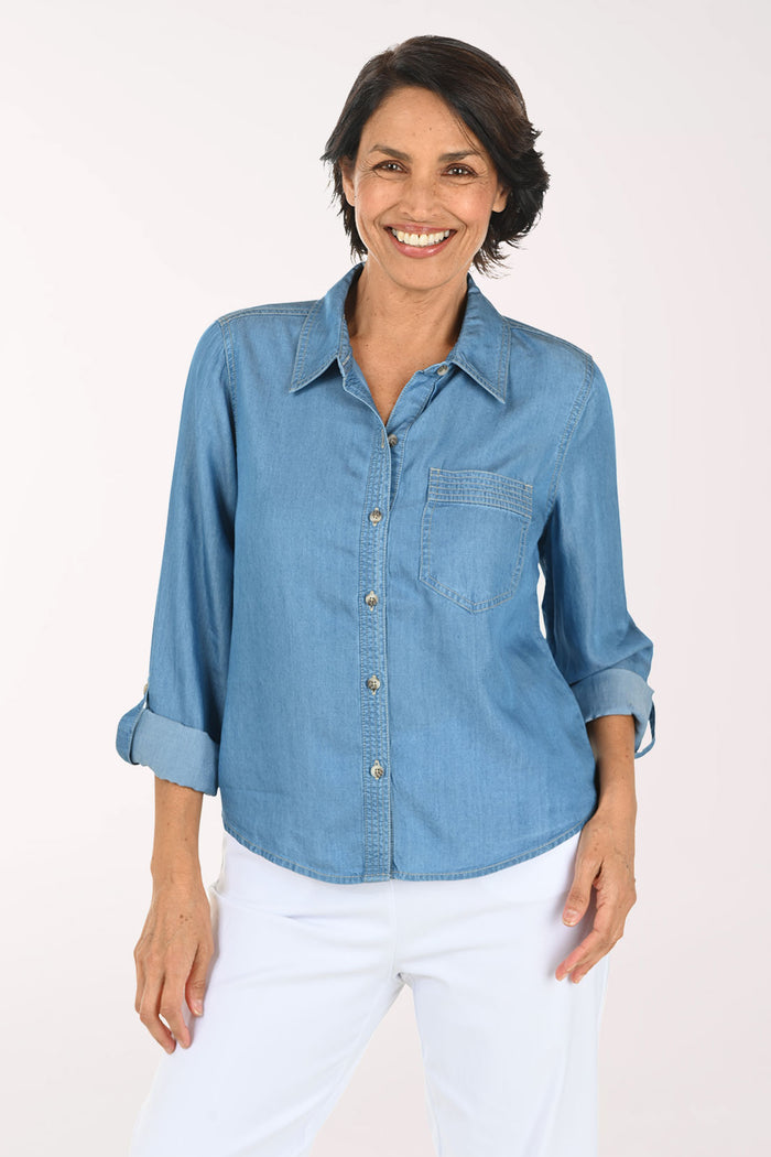 A smiling woman wearing a light blue chambray button-down shirt with rolled-up sleeves and white pants, standing against a plain off-white studio background.