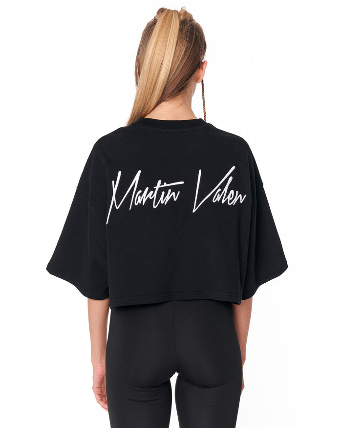 A woman shown from the back wearing a black cropped t-shirt with 'Martin Valen' printed in white script across the back, paired with black leggings, against a plain white background.