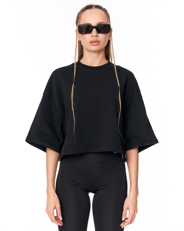 A model wearing a black oversized short-sleeve crop top with tonal graphic embroidery, paired with black leggings and black sunglasses, against a plain white background.