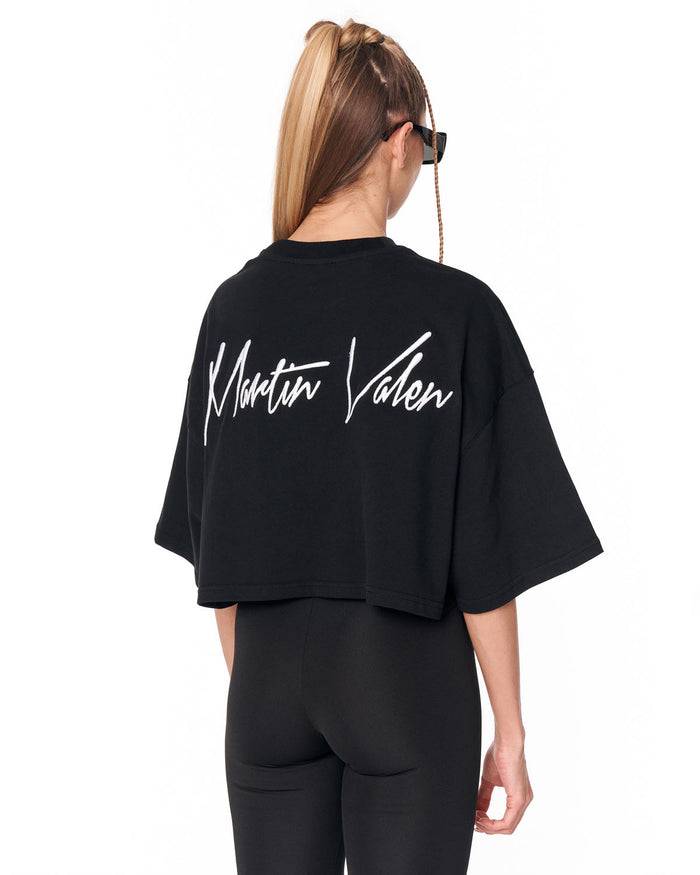 A woman shown from the back wearing a black cropped T-shirt with a large white 'Martin Valen' script logo across the back, paired with black leggings, against a plain white background.