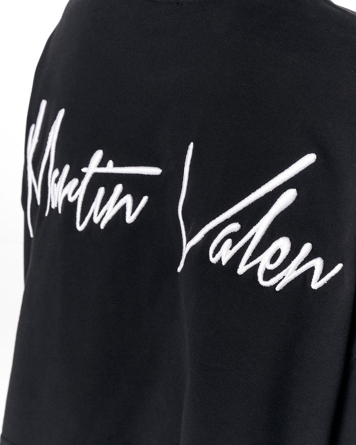 Close-up of a black sweatshirt featuring a large white embroidered script logo that reads 'Martin Valen' across the chest.
