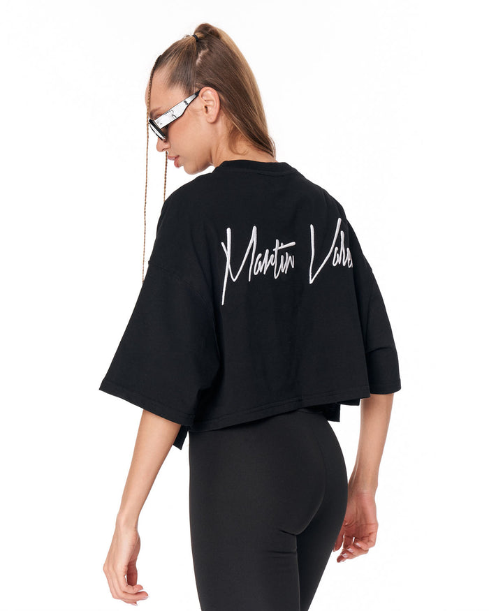 A woman shown from the back wearing a black cropped t-shirt with 'Maison Louis Vuitton' printed in white script across the upper back, paired with black leggings, against a plain white background.