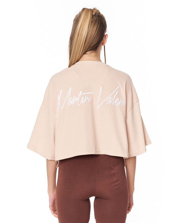 A woman shown from the back wearing a light beige cropped t-shirt with 'Martin Valen' printed in white script across the back, paired with brown leggings, against a plain white background.