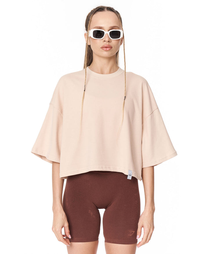 A model wearing a light beige oversized short-sleeve crop top and dark brown bike shorts, styled with white-framed sunglasses, against a plain white background.
