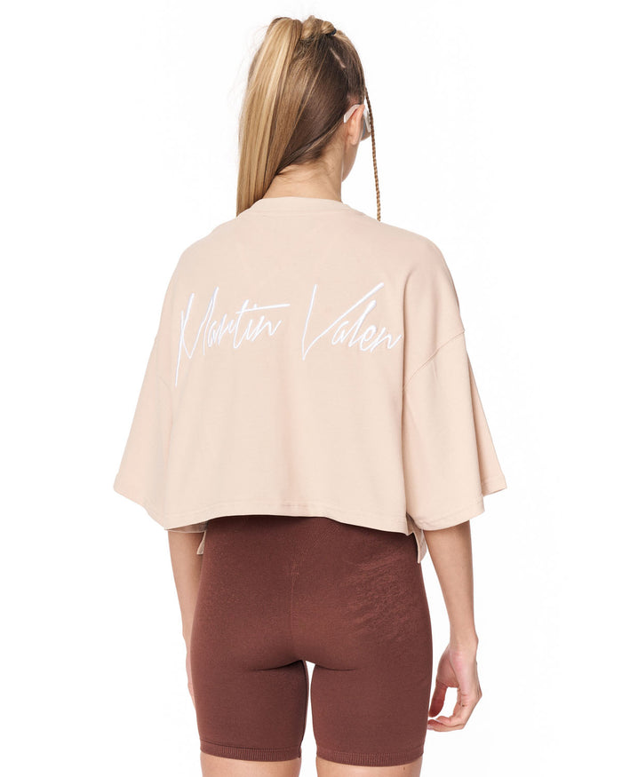 A woman shown from the back wearing a light beige cropped t-shirt with 'Martin Valen' written in white script across the back, paired with brown bike shorts, against a plain white background.