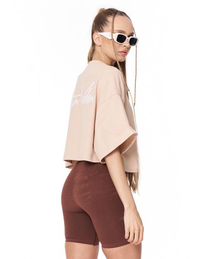 A woman shown from the back wearing a light beige oversized crop top with white script text on the back and dark brown fitted biker shorts, against a plain white background.