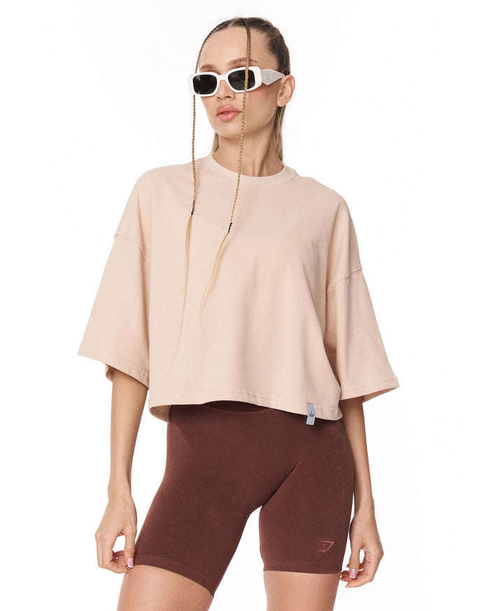 A model wearing a light beige oversized short-sleeve crop top and dark brown fitted bike shorts, styled with white sunglasses, against a plain white background.