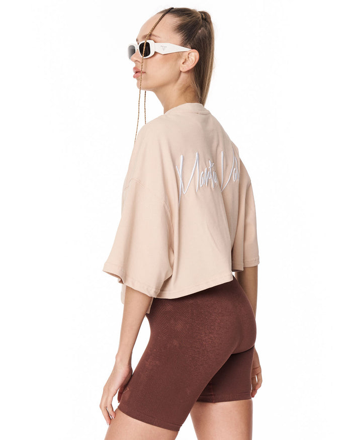 A woman shown from the back wearing a beige cropped t-shirt with white script lettering on the back, paired with brown bike shorts, against a plain white background.