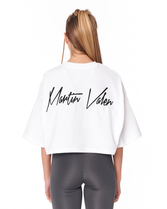 A woman viewed from behind wearing a white cropped T-shirt with 'Martin Valen' printed in black script across the back, paired with dark gray leggings, against a plain white background.