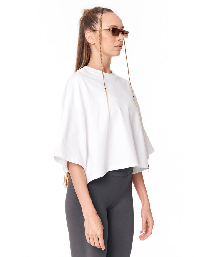 A woman in profile wearing a white short-sleeve poncho-style top, dark gray leggings, and gold chain-link sunglasses with long dangling chains, against a plain white background.