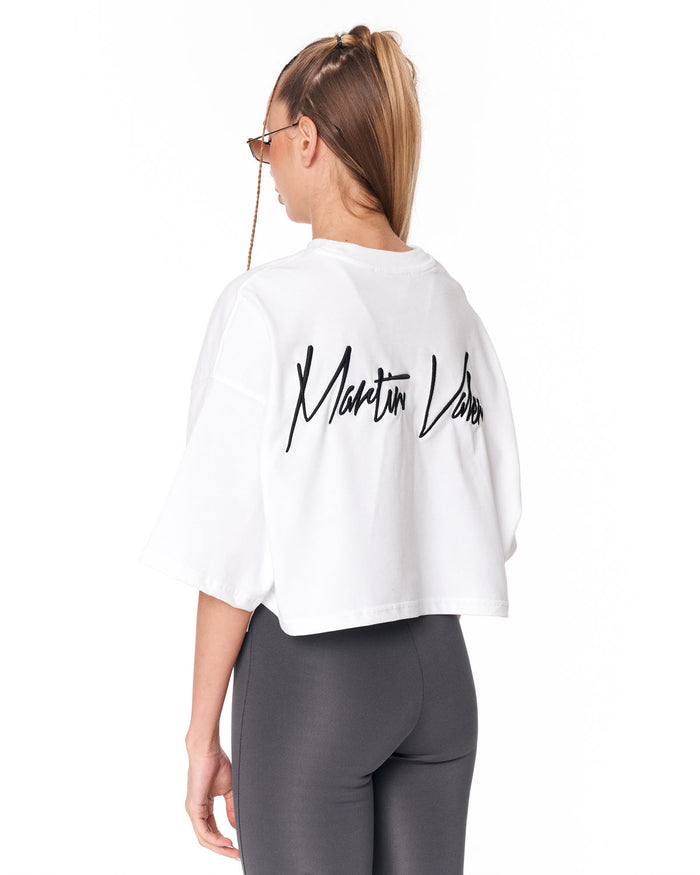 A woman shown from the back wearing a white cropped t-shirt with a black script logo on the back, paired with dark gray leggings, against a plain white background.