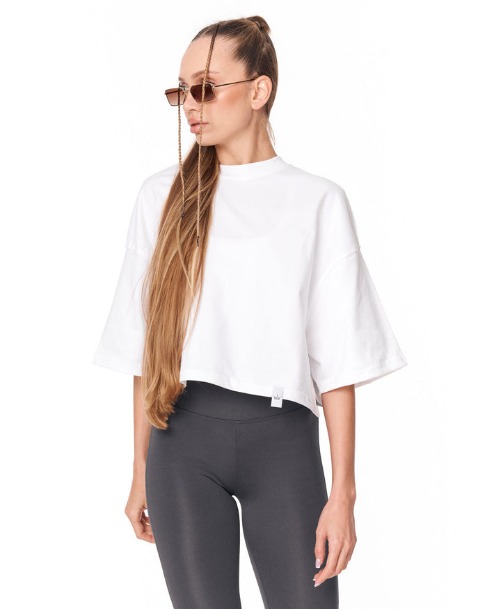 A woman modeling a white short-sleeve crop top with a high mock neck and dark gray leggings against a plain white background.