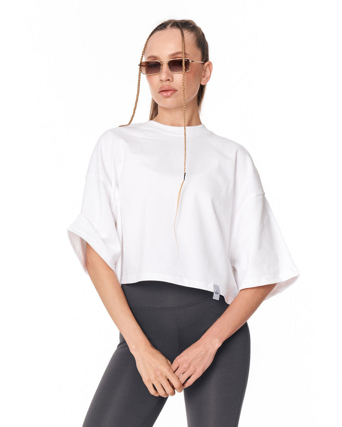 A woman modeling a white cropped tunic top with wide, draped sleeves and a front zipper, paired with dark gray leggings, against a plain white background.