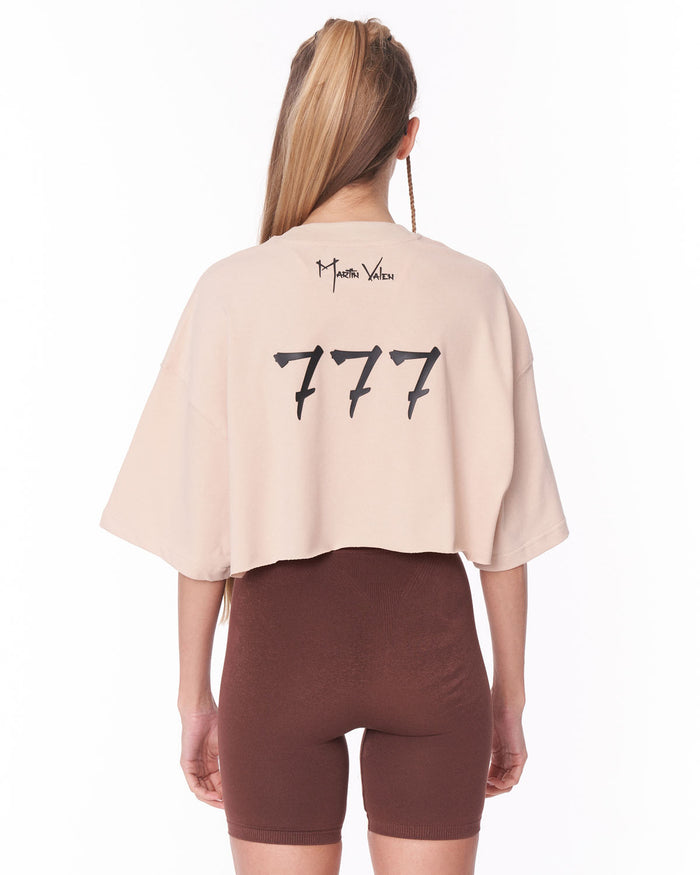 A woman shown from the back wearing a light beige cropped t-shirt with 'Martin Valen' and '777' printed in black, paired with brown bike shorts, against a plain white background.