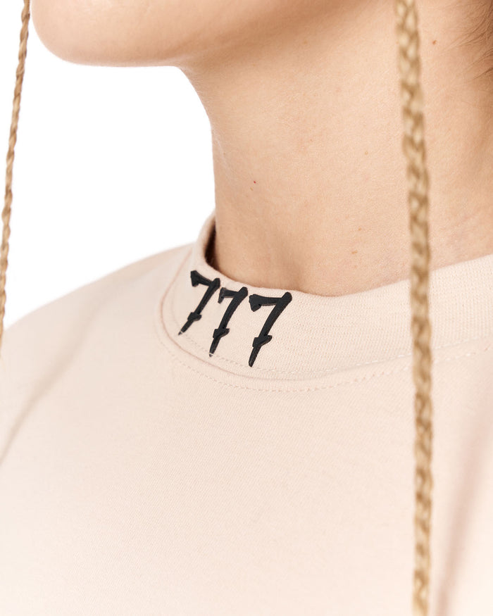 Close-up of a person wearing a light beige crewneck sweatshirt with a black embroidered '777' logo on the collar, featuring a gold chain necklace hanging down.