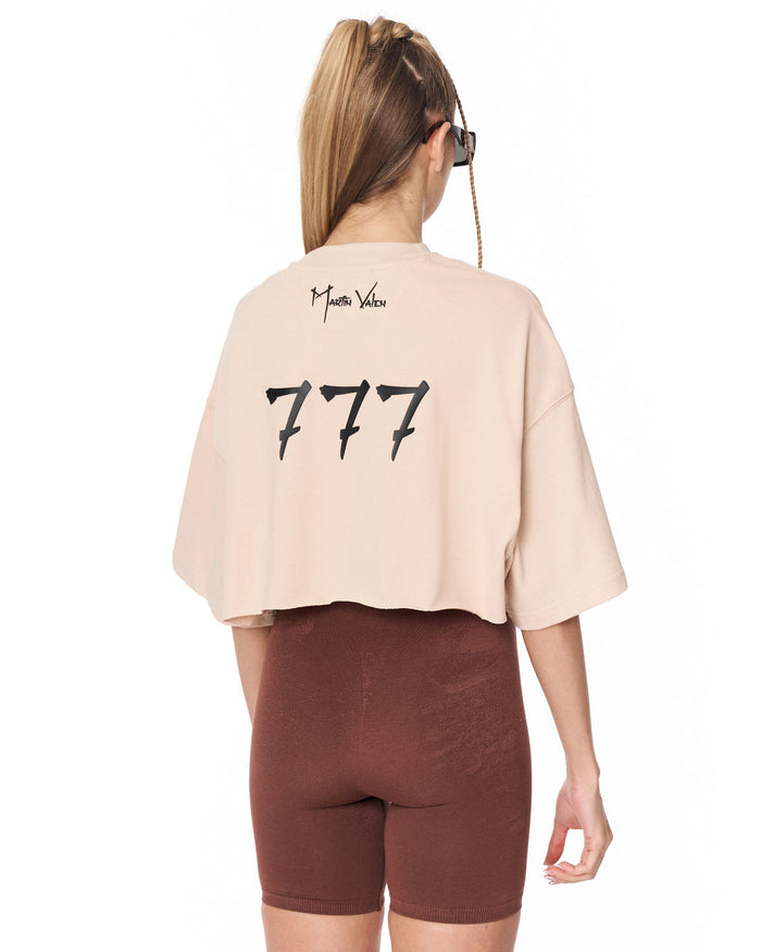 A woman shown from the back wearing a light beige cropped t-shirt with 'Martin Valen' and '777' printed on the back, paired with brown fitted shorts against a white background.