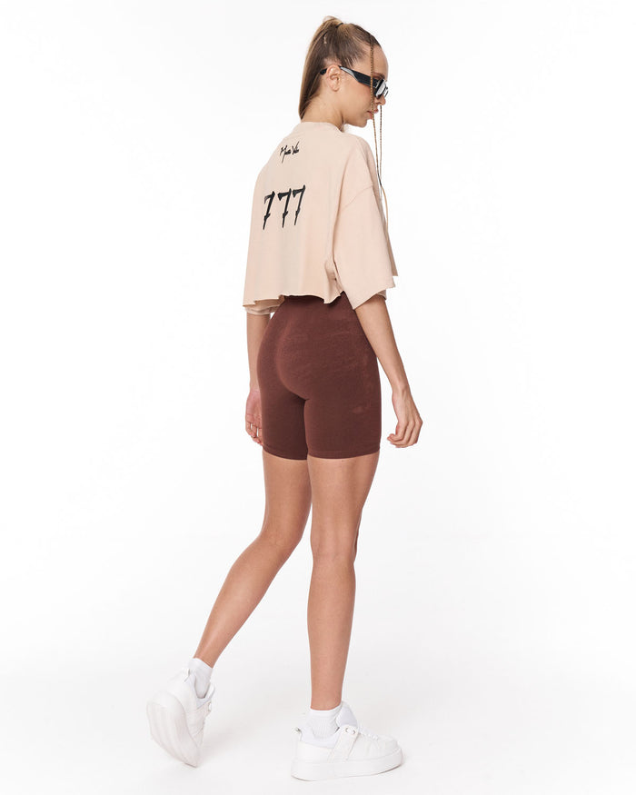 A model shown from the back wearing a light beige cropped t-shirt with black graphic text and dark brown fitted biker shorts, paired with white sneakers, against a plain white studio background.