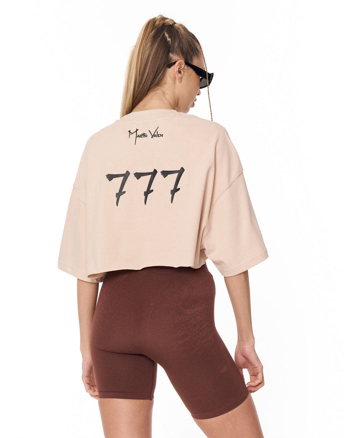 A woman shown from the back wearing a cropped beige t-shirt with a black '777' graphic and 'Martin Valen' logo, paired with brown fitted shorts against a white background.