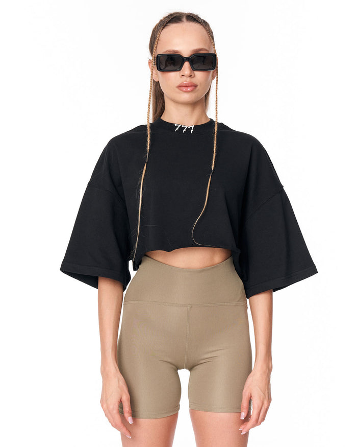 A model wearing a black cropped short-sleeve top with wide sleeves and beige high-waisted biker shorts, styled with black sunglasses and a silver necklace against a plain white background.