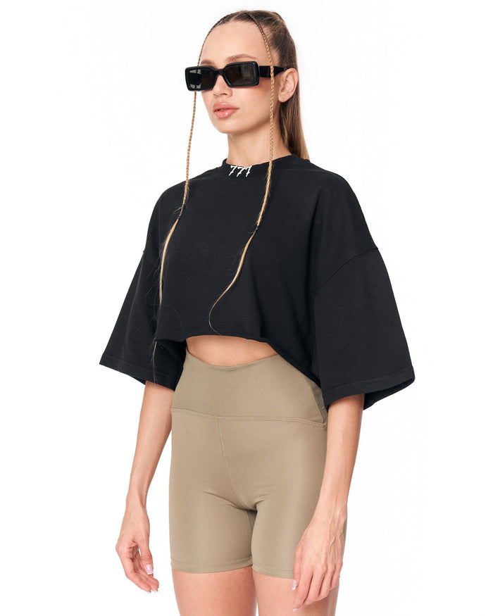 A model wearing a black short-sleeve crop top with wide bell sleeves and beige high-waisted bike shorts, styled with black sunglasses and a gold chain necklace against a plain white background.