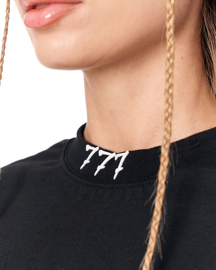 Close-up of a person wearing a black crewneck sweatshirt with a white embroidered '777' logo on the collar, featuring blonde braided hair against a white background.