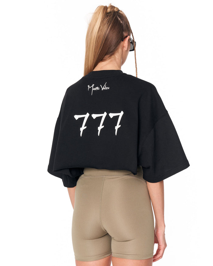 A woman shown from the back wearing an oversized black cropped T-shirt with a large white '777' graphic and 'Mia Mia Vito' text on the back, paired with beige fitted shorts against a plain white background.