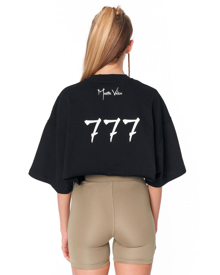 A woman shown from the back wearing a black oversized crop top with white '777' text and a small 'Martin Valen' signature graphic, paired with beige fitted shorts, against a plain white background.