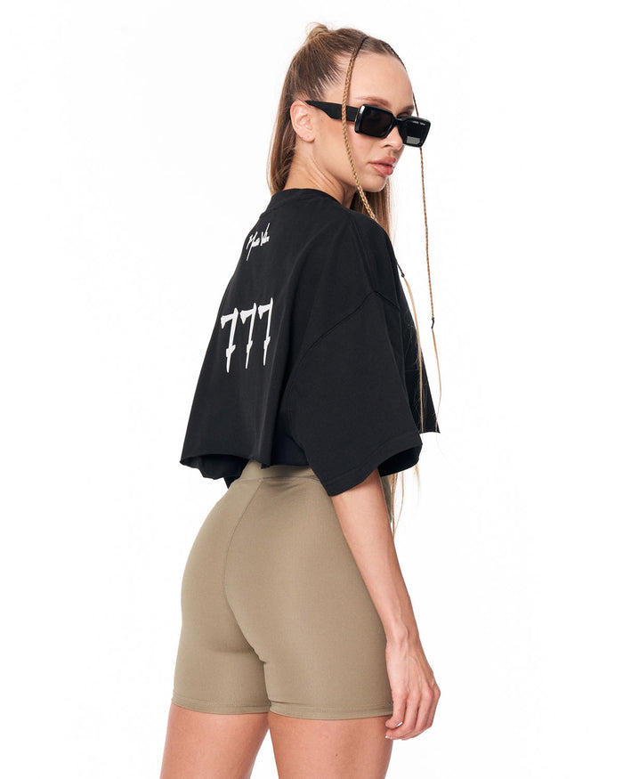 A woman shown from the back wearing a black oversized cropped T-shirt with white graphic text and tan fitted shorts against a plain white studio background.