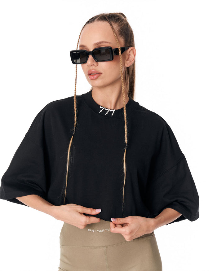 A woman modeling a black oversized short-sleeve top with a crew neck, paired with beige pants and black sunglasses with braided chains, against a plain white background.