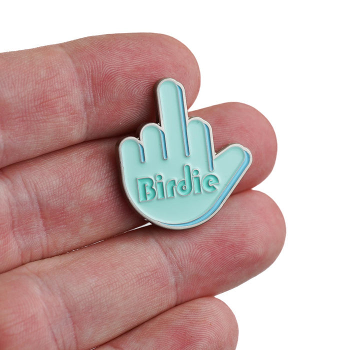 A hand holds the Birdie Finger - Ball Marker, a light blue enamel pin shaped like a hand giving the middle finger with "Birdie" embossed on the palm. Iron nickel plating adds boldness, while a blurred background makes it stand out.