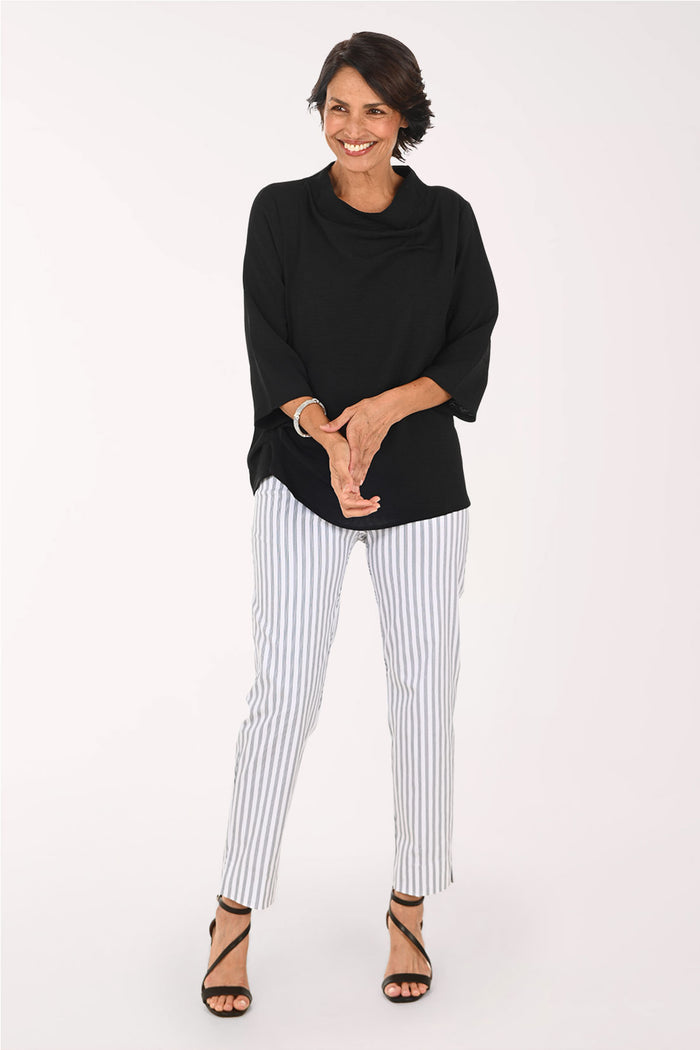 A woman modeling a black cowl-neck top and white pants with thin blue vertical stripes, paired with black strappy high-heeled sandals, against a plain white studio background.