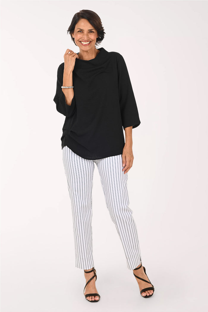 A woman modeling a black draped three-quarter sleeve top and white pants with thin black vertical stripes, paired with black strappy heels, against a plain white studio background.
