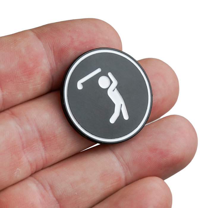 A hand holds the Bogeyman - Ball Marker, a round, black metal token with a white stick figure golfer. It has a smooth, glossy finish with iron nickel plating and a thin white border around the edge.