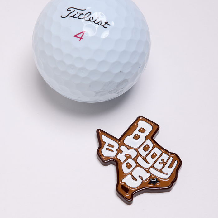 A white Titleist 4 golf ball sits above a Texas-shaped brown and white keychain that reads "Bogey Bros" in large, playful letters, complete with a small black star in the lower left corner. Nearby, the Burnt Ends - Ball Marker, crafted with iron nickel plating for a striking shine, completes the arrangement—all displayed on a plain light surface.