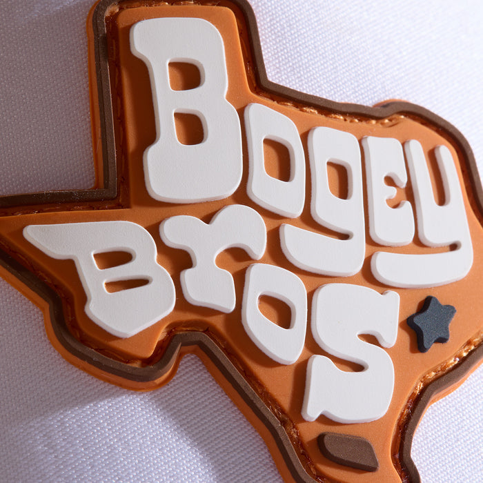 An orange Texas-shaped patch with bold white letters reads Bogey Bros. A small dark star marks the lower-right area. The patch is on a white fabric background.