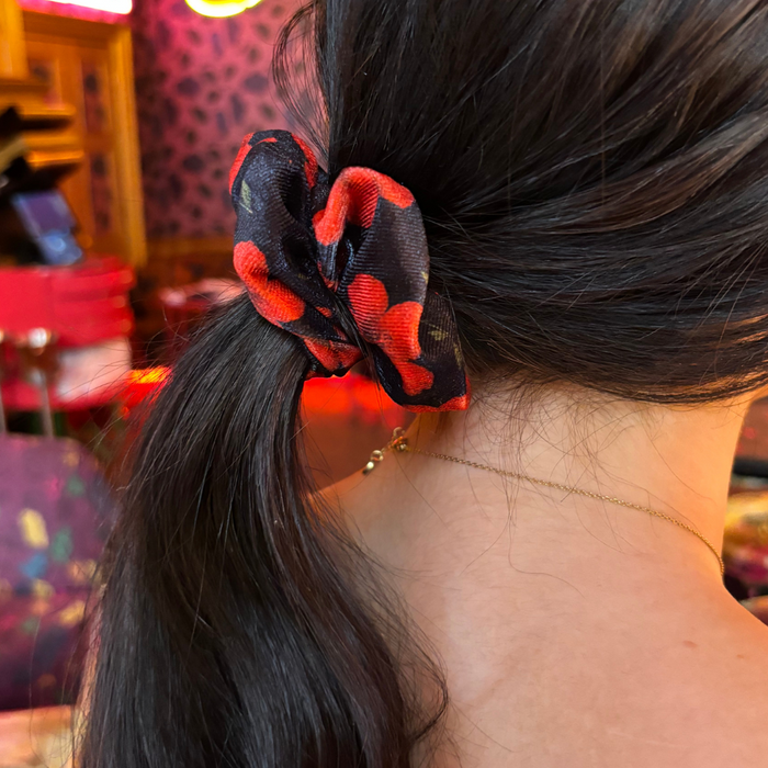 Dark-haired person wearing a cherry-patterned hibiscus scrunchie in their low ponytail, shown in warm light.