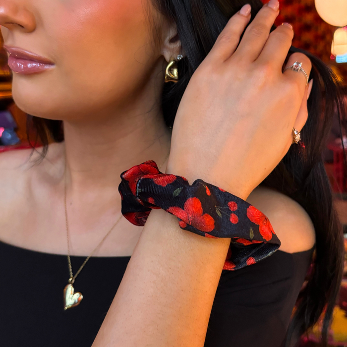 Woman wearing a black top and gold jewelry adjusts her hair with a red Cherry Hibiscus patterned scrunchie.