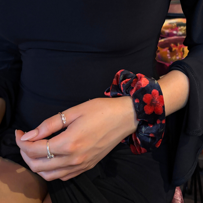 Person in black with hands on lap, wearing rings and a Coconut Lane scrunchie with red cherry and hibiscus print.