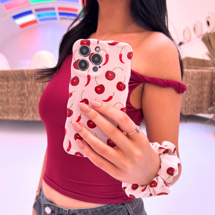 Woman in maroon top takes mirror selfie wearing a red scrunchie with cherry print, holding her phone.